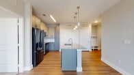 This well-appointed single-floor residence showcases a thoughtfully designed open layout with a seamless flow between living and kitchen areas. The kitchen serves as the centerpiece, featuring a large central island topped with granite countertops and an integrated sink with a brushed gold faucet, illuminated by elegant pendant lighting with brass accents. Two-tone cabinetry — soft gray on the lower cabinets and cream-white on the uppers — pairs with a white tile backsplash and a full suite of black appliances, including a French door refrigerator, range, and over-the-range microwave. Warm wood-look hardwood flooring runs throughout the main living areas, adding continuity and warmth. A built-in mudroom-style nook near the entry provides convenient storage. The bathroom offers a soaking tub with large-format tile surround, a granite-topped vanity with a dark blue cabinet, and wood-look flooring. Adjacent to the bathroom, a spacious walk-in closet with wire shelving provides ample storage. The carpeted bedroom receives generous natural light through a pair of large windows with exterior views, creating a bright and comfortable retreat.
