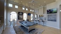 This single-floor entertainment space showcases a thoughtfully designed open-concept layout that seamlessly blends recreation and hospitality. The room is anchored by a sleek, contemporary pool table with a grey felt surface set atop a plush area rug, creating a defined game zone within the expansive space. Soaring ceilings with clerestory windows along the upper walls flood the interior with natural light, while recessed lighting and dual ceiling fans ensure comfortable illumination and airflow throughout. A series of elegant arched windows along one wall frames outdoor views and reinforces the room's sophisticated architectural character. The wet bar area is finished with white cabinetry, a contrasting tile backsplash, a stainless steel range hood, and modern bar seating, making it well-suited for entertaining. Multiple wall-mounted flat-screen televisions are strategically positioned for viewing from various areas of the room. Light hardwood flooring runs throughout, complementing the neutral, warm-toned walls and contributing to the bright, airy ambiance of the space. The overall design aesthetic is clean, modern, and highly functional.