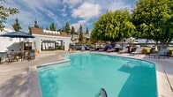 Located at 140 S Highland Ave in Placentia, California, this single-story community space offers a welcoming outdoor environment designed for leisure and social gatherings. The centerpiece of the property is a generously sized swimming pool with a classic freeform shape, featuring clear turquoise water and a wading step entry. Surrounding the pool is a well-maintained concrete deck outfitted with multiple lounge chairs, providing ample space for sunbathing and relaxation under the Southern California sun. A charming clubhouse structure anchors one side of the pool area, finished with light gray siding, a covered patio, and skylights that allow natural light into the interior. Outdoor seating and tables are visible beneath the covered patio, offering a shaded gathering spot. Mature trees frame the space, including a large, lush canopy tree that provides natural shade and enhances the park-like ambiance. The fenced perimeter ensures privacy and security while maintaining an open, airy feel. The surrounding hillside residential setting and abundant greenery contribute to a serene, resort-like atmosphere. This community amenity space is thoughtfully designed to encourage outdoor activity and relaxation in a pleasant, well-landscaped environment.