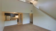 This two-story loft-style space is defined by its dramatic double-height ceilings in the main living area, creating an expansive, airy atmosphere. The living area is finished with neutral carpet flooring and smooth, light-colored walls that enhance the sense of openness. A staircase runs along one side of the space, leading to the upper level, which overlooks the main floor via an open mezzanine railing — a defining architectural feature that adds depth and visual interest to the interior.

The kitchen is integrated into the main floor and features dark cabinetry contrasted against lighter countertops and backsplash tile, along with under-cabinet lighting that provides a warm, functional glow. A peninsula counter separates the kitchen from the living area, offering a natural transition between the two spaces. A doorway near the kitchen provides access to an additional room or utility area.

The upper level, visible from below, benefits from recessed lighting and appears to serve as a secondary functional space overlooking the living area. Overall, the layout is open and practical, with the double-height design serving as the standout architectural element throughout.