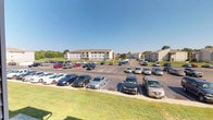 This residential complex presents a well-organized, single-level-access layout surrounded by an expansive outdoor environment. The exterior view reveals multiple mid-rise residential buildings, each featuring a neutral facade with clean architectural lines and private balconies that offer residents outdoor space and open views. The buildings are arranged to create a cohesive community setting, with generous paved parking areas that provide convenient, direct access for residents and guests alike. The parking areas are clearly marked and spacious, accommodating a large number of vehicles with ease. Lush green lawns border the parking and walkway areas, adding a natural, park-like quality to the grounds. Mature trees are visible in the background, contributing to a sense of privacy and a connection to the surrounding landscape. The complex benefits from abundant natural light, with the open layout allowing sunlight to reach both the building facades and the outdoor common areas throughout the day. The overall ambiance is functional yet inviting, with a well-maintained exterior that reflects a commitment to upkeep and community living. The open sky and green surroundings enhance the sense of space, making the outdoor areas feel expansive and welcoming.