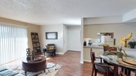 This single-floor residence showcases an open-concept design that seamlessly integrates the living, dining, and kitchen areas into one cohesive space. The warm hardwood flooring flows continuously throughout, lending a sense of unity and warmth across the main living areas.

The living room is a comfortable, well-lit space featuring a textured ceiling and neutral beige walls that create a versatile backdrop for furnishings. A leaning bookshelf adds functional storage and decorative character, while a patterned area rug anchors the seating arrangement.

Adjacent to the living area, a dining space accommodates a glass-top table and chairs, positioned conveniently close to the kitchen. The kitchen is accessible via a half-wall counter, which serves as both a visual divider and a casual breakfast bar. White appliances, including a range, are visible within the kitchen space.

Near the entrance, a set of double closet doors provides generous storage directly off the main living area. Natural light filters into the space through windows, contributing to the bright and welcoming ambiance throughout this efficiently designed, single-level home.