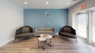 Located at 735 Randolph St in Canton, MA, Blue Hills Village offers a thoughtfully designed single-floor community space built for comfort and social gathering. The interior is finished with light wood-look flooring throughout, creating a warm and cohesive aesthetic across all areas.

The lounge area serves as a central gathering point, furnished with a neutral sofa and a pair of dark leather accent chairs arranged around geometric marble-top coffee tables. A calming blue accent wall anchors the seating area, providing a distinct visual focal point against the soft gray walls found throughout the space.

Adjacent to the lounge, a dedicated dining area features multiple rectangular tables paired with light blue chairs, offering ample seating for group meals or community events. The "Blue Hills Village" branding is prominently displayed on the wall, reinforcing the sense of place and community identity.

The kitchen is well-appointed with white shaker-style cabinetry, dark laminate countertops, a stainless steel full-size refrigerator, a microwave, a coffee maker, and a utility sink — providing all the essentials for resident use and event preparation.

Natural light filters in through frosted glass entry doors in the lobby-facing area, which also includes additional casual seating. Recessed lighting throughout ensures the space remains bright and inviting at all times.
