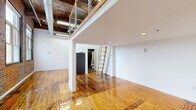 This two-floor loft unit at 7 Rowe Ave in Hartford, Connecticut, blends industrial character with modern functionality. The main floor serves as the primary living area, featuring polished hardwood floors, soaring ceilings, and exposed ductwork that highlight the space's industrial roots. Striking exposed brick walls and large multi-pane windows flood the area with natural light while offering views of the surrounding cityscape. A compact, well-appointed kitchen occupies one corner, outfitted with white cabinetry, dark granite countertops, and a full suite of stainless steel appliances including a gas range, dishwasher, microwave, and refrigerator. The bathroom is finished with large-format gray tile throughout, featuring an open walk-in shower with a curtain rod and modern fixtures. A wooden ladder staircase leads to the mezzanine level, secured by a cable railing system, which provides additional flexible living or sleeping space with its own closet. The upper level continues the hardwood flooring and benefits from recessed lighting. The overall ambiance is that of a classic urban loft — open, airy, and rich with architectural detail that balances raw industrial elements with clean, contemporary finishes.