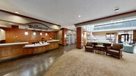 The Wellington, located at 2235 Rockwood Ave in Saint Paul, Minnesota, is a well-appointed senior living community offering nearly 6,000 square feet of thoughtfully designed common space on a single floor. The welcoming main entrance opens into a grand lobby anchored by a curved wooden reception desk prominently branded with The Wellington name. Rich hardwood-style flooring transitions into patterned carpeting, setting a warm and refined tone throughout.

The central atrium is a standout architectural feature, rising to a dramatic two-story height with upper-level balcony railings and large windows that flood the space with abundant natural light. Multiple seating areas are distributed throughout, including cozy nooks with armchairs positioned beside floor-to-ceiling windows overlooking manicured landscaping. A tiled fireplace with decorative ironwork wall art serves as an inviting focal point in one of the lounges.

A spacious dining and activity area features a distinctive wood-planked ceiling with pendant drum lighting, combining warmth and modern elegance. Elevator access and French-style glass-paned doors connect the various wings. Throughout the facility, neutral tones, crown molding, wood accents, and decorative columns contribute to a cohesive, upscale ambiance designed for comfort and community.