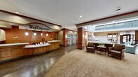 The Wellington, located at 2235 Rockwood Ave in Saint Paul, Minnesota, is a well-appointed senior living community offering nearly 6,000 square feet of thoughtfully designed common space on a single floor. The welcoming main entrance opens into a grand lobby anchored by a curved wooden reception desk prominently branded with The Wellington name. Rich hardwood-style flooring transitions into patterned carpeting, setting a warm and refined tone throughout.

The central atrium is a standout architectural feature, rising to a dramatic two-story height with upper-level balcony railings and large windows that flood the space with abundant natural light. Multiple seating areas are distributed throughout, including cozy nooks with armchairs positioned beside floor-to-ceiling windows overlooking manicured landscaping. A tiled fireplace with decorative ironwork wall art serves as an inviting focal point in one of the lounges.

A spacious dining and activity area features a distinctive wood-planked ceiling with pendant drum lighting, combining warmth and modern elegance. Elevator access and French-style glass-paned doors connect the various wings. Throughout the facility, neutral tones, crown molding, wood accents, and decorative columns contribute to a cohesive, upscale ambiance designed for comfort and community.