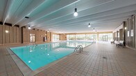 Located on Wooster Road in Rocky River, Ohio, this single-floor recreation facility is thoughtfully designed to support a wide range of wellness and leisure activities. The centerpiece of the space is a large indoor lap pool, enclosed within a generously proportioned natatorium featuring warm wood-paneled walls, a beamed ceiling, and extensive floor-to-ceiling windows that flood the area with natural light and offer views of the surrounding greenery. Tiled pool decking lines the perimeter, providing ample space for swimmers and observers alike. A hot tub is also present, situated adjacent to the fitness center area. The fitness center is well-equipped with cardio machines and exercise equipment, separated from the pool deck by a railed partition. Wide tiled corridors connect the various zones of the facility, lending a cohesive, campus-like feel to the interior. A bright, open common area with large glass walls and direct access to an outdoor patio provides a welcoming gathering space filled with natural light. The outdoor area includes seating and additional recreation space surrounded by mature hedging. Throughout, the architecture blends functional design with warm materials, creating an inviting and active environment for residents and members of the Rocky River community.