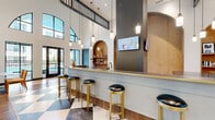 This thoughtfully designed single-floor amenity space combines contemporary elegance with functional versatility across several distinct zones. The centerpiece is a dramatic double-height lobby area, where soaring ceilings, arched transom windows, and bold checkered tile flooring create a striking first impression. Warm hardwood flooring transitions throughout the remaining areas, tying the spaces together cohesively.

A sleek bar counter with gold-trimmed edges and black leather-topped brass stools anchors one side of the main hall, complemented by pendant lighting and a built-in arched display niche. Large windows and glass doors flood the space with natural light and offer views of an outdoor amenity area. A lounge corner features a fireplace, mounted television, and plush seating beneath a tray ceiling with bold blue accents.

Intimate arched lounge nooks provide tufted emerald green banquette seating with marble-topped tables, ideal for casual gatherings. The conference room accommodates a full-length wooden table beneath a statement crystal chandelier, with built-in cabinetry and a view into an adjoining lounge. A dedicated business center offers a print station, dark marble cabinetry, wood upper cabinets, and ample counter workspace, completing the suite of professional amenities.