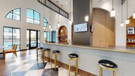 This thoughtfully designed single-floor amenity space combines contemporary elegance with functional versatility across several distinct zones. The centerpiece is a dramatic double-height lobby area, where soaring ceilings, arched transom windows, and bold checkered tile flooring create a striking first impression. Warm hardwood flooring transitions throughout the remaining areas, tying the spaces together cohesively.

A sleek bar counter with gold-trimmed edges and black leather-topped brass stools anchors one side of the main hall, complemented by pendant lighting and a built-in arched display niche. Large windows and glass doors flood the space with natural light and offer views of an outdoor amenity area. A lounge corner features a fireplace, mounted television, and plush seating beneath a tray ceiling with bold blue accents.

Intimate arched lounge nooks provide tufted emerald green banquette seating with marble-topped tables, ideal for casual gatherings. The conference room accommodates a full-length wooden table beneath a statement crystal chandelier, with built-in cabinetry and a view into an adjoining lounge. A dedicated business center offers a print station, dark marble cabinetry, wood upper cabinets, and ample counter workspace, completing the suite of professional amenities.