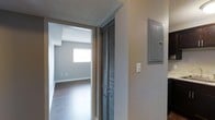 This single-floor space is captured through a hallway that serves as a functional transition between areas. The corridor features neutral grey walls and a textured ceiling, lending a clean, understated aesthetic throughout. A set of louvered bi-fold closet doors lines one side of the hallway, providing practical storage while adding a classic architectural detail. Mounted on the opposite wall is a residential electrical panel, indicating convenient utility access within the living space. The hallway leads into an adjoining room that benefits from natural light streaming through a window, illuminating the space and highlighting the hardwood-style flooring that runs throughout. The room beyond appears open and unfurnished, offering a clear view of its layout and potential. The overall color palette — composed of soft greys and warm wood tones — creates a cohesive, modern feel. The straightforward design prioritizes functionality while maintaining a tidy, well-maintained appearance, making this single-floor layout a practical and versatile living environment.