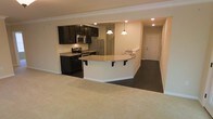 This single-floor residence showcases an open-concept layout that seamlessly connects the kitchen and living area, creating a spacious and functional environment. The kitchen is outfitted with dark espresso cabinetry that provides strong visual contrast against the light-toned walls and countertops. Stainless steel appliances, including a refrigerator, microwave, and range, contribute a modern, polished aesthetic. A curved peninsula-style island with a light stone countertop extends into the living space, featuring decorative corbel brackets and bar-height seating. Two pendant lights hang above the island, providing focused task lighting that complements the recessed ceiling lights distributed throughout. The kitchen features dark tile flooring, which transitions into plush, neutral-toned carpet in the living area, clearly delineating the two zones while maintaining visual cohesion. Crown molding along the ceiling adds a refined architectural detail throughout the open area. A hallway visible in the background leads to additional rooms, hinting at further living space beyond the main area. Overall, the space presents a well-balanced combination of practical design and contemporary finishes.