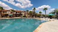 This outdoor pool area is designed to evoke a resort-style atmosphere, centered around a generously sized swimming pool with clear, blue-green water. The pool deck is finished in light-toned concrete, providing ample space for sunbathing and relaxation. Multiple lounge chairs are arranged around the pool, offering both open sun exposure and shaded retreats beneath teal market umbrellas and a wood-beam pergola structure. A spa is positioned adjacent to the main pool, adding an additional amenity to the space.

A single-story clubhouse building anchors one side of the pool area, featuring a stucco exterior, a tile roof with Mediterranean-inspired detailing, and large glass sliding doors that open directly onto the pool deck. The building's entrance is framed by dark wood columns and accents, with wall-mounted lighting and posted pool rules signage nearby. Tall palm trees and mature shade trees are interspersed throughout the surrounding landscape, providing natural screening and contributing to the tropical ambiance. The multi-story residential buildings surrounding the pool are visible from the deck, with some units offering direct views of the pool through sliding glass doors and balconies.