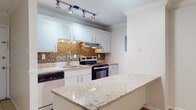 Located on Village Lane in Winter Park, Florida, this single-story residence offers a thoughtfully arranged layout with clean, bright interiors throughout. The kitchen is a standout feature, equipped with white shaker-style cabinetry, granite countertops, stainless steel appliances including an electric range and dishwasher, and a mosaic tile backsplash that adds visual texture. Track lighting illuminates the workspace, and a peninsula counter provides additional prep space while creating an open connection to the adjacent dining area.

The living area is generously proportioned, featuring light wood-style flooring, crown molding, and a ceiling fan overhead. Large sliding glass doors with vertical blinds open to an outdoor space, allowing natural light to flow into the room. A wall-mounted air conditioning unit serves the living space, and a second window adds additional ventilation.

The bathroom is cleanly finished with tile flooring, a white vanity with granite countertop, and a full tub-shower combination with white tile surround. The overall interior palette is neutral and bright, with white walls and crown molding maintaining a cohesive, polished feel throughout the home.