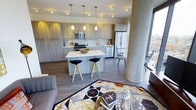This single-floor residence showcases a thoughtfully designed open-concept layout that seamlessly connects the kitchen and living areas, creating a spacious and cohesive environment. The kitchen is a standout feature, outfitted with flat-panel cabinetry in a warm wood-tone finish, stainless steel appliances including a gas range, microwave, and a large French-door refrigerator, and a crisp white quartz island with seating for two modern bar stools. Pendant lighting above the island adds a refined touch, while track lighting throughout ensures the space is well-illuminated. Wide-plank wood-tone flooring runs throughout the open floor plan, anchoring the space with warmth and continuity. A distinctive structural column with a smooth cylindrical finish adds an architectural element to the living area, where a plush sofa and a bold geometric-patterned area rug define the lounge space. Floor-to-ceiling windows allow abundant natural light to pour in, offering expansive city views that elevate the overall ambiance. The combination of modern finishes, an efficient layout, and striking urban vistas makes this a well-appointed and visually compelling living space.