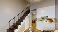 This two-story loft offers a thoughtfully designed living experience that blends industrial character with modern comfort. The main level features an open-concept layout where the living area flows seamlessly into the kitchen and dining space. The kitchen is outfitted with white cabinetry, a functional island or breakfast bar, and standard appliances, providing both style and practicality. The dining area sits just adjacent, making the space ideal for everyday living and entertaining. A standout architectural feature is the staircase, which showcases rich dark hardwood treads set against crisp white risers and framed by a sleek black metal railing — a visually striking element that anchors the space. The staircase leads to the upper level, which hosts the bedroom loft area, offering a sense of privacy and separation from the main living floor. Warm hardwood flooring runs throughout, adding continuity and a natural, inviting feel to the space. The high ceilings characteristic of loft-style living create an airy, open atmosphere, with visible HVAC elements above reinforcing the industrial aesthetic. Neutral wall tones throughout allow the architectural details to take center stage, resulting in a cohesive and versatile living environment.