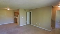This two-floor residential space offers a functional and straightforward layout designed for comfortable everyday living. The main living area is finished with wall-to-wall carpeting in a neutral tone, providing a warm and understated foundation that complements the beige-painted walls running throughout. A textured popcorn ceiling adds a classic touch overhead, while a central HVAC vent ensures climate comfort year-round. White baseboards frame the perimeter of the room, lending a clean, finished look to the space. An open passageway connects the living area to the kitchen, which features warm wood cabinetry, a white range, and dark countertops — offering a practical cooking environment. The kitchen's lighter flooring contrasts neatly with the carpeted living space, providing a subtle but effective visual separation between the two areas. A doorway from the main room leads to additional spaces within the home, hinting at the multi-room layout spread across two floors. The overall ambiance is clean, neutral, and move-in ready, making it well-suited for those looking for a no-frills, comfortable living environment with a straightforward and efficient use of space.