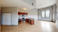Located at 1140 E. Market St in Akron, Ohio, this two-bedroom, two-bathroom loft-style unit offers an open, airy atmosphere defined by soaring ceilings, exposed white-painted brick walls, and light hardwood flooring throughout the main living areas. The open-concept layout seamlessly connects the living room, dining area, and kitchen, creating a generous and flexible living space. A bank of large, industrial-style windows floods the interior with natural light and frames scenic outdoor views of the surrounding Akron landscape. Ceiling fans are installed throughout, adding both function and style. The kitchen features rich cherry wood cabinetry, stainless steel appliances, a tile backsplash, and a peninsula with pendant lighting that anchors the space. Both bedrooms are carpeted and receive ample natural light through oversized windows with roller shades, and the primary bedroom is notably spacious. The unit includes four closets, one of which is a walk-in with wire shelving. The bathrooms are clean and modern, with one featuring a walk-in shower, wood-tone vanity, and tile flooring. A well-lit hallway connects the living areas to the private quarters, completing this distinctive urban retreat.