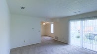 This single-floor residence offers a clean, open layout with a straightforward and functional design. The living area is generously proportioned, featuring neutral-toned carpet that extends throughout the space, creating a cohesive and understated aesthetic. Smooth, light-colored walls are complemented by a textured ceiling, adding subtle dimension to the interior. A sliding glass door floods the living space with natural light and provides direct access to an outdoor area, while vertical blinds offer privacy and light control. An open passageway connects the living area to an adjoining dining space, which is accented by a pendant light fixture that adds a modest decorative touch. A window in the dining area further enhances the natural brightness of the space. HVAC vents are positioned throughout the floor, ensuring efficient climate control. The overall ambiance is bright, neutral, and move-in ready, offering a versatile canvas well-suited for a variety of interior styles. The single-floor layout provides ease of navigation and accessibility throughout the home.