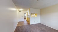 This single-story unit at 601–621 N Hayden Dr in Tucson, Arizona offers a straightforward, functional layout connecting several well-defined living spaces. The living area is finished with wall-to-wall carpet and features neutral-toned walls beneath a textured ceiling, creating a quiet, understated atmosphere. A pass-through window connects the living space directly to the kitchen, maintaining visual openness between the two areas.

The kitchen is a galley-style space laid with wood-look plank flooring and lined with warm-toned wood cabinetry above and below the counters. It includes a range with an overhead exhaust hood, a full-size refrigerator, and a dishwasher, providing essential appliances within a compact, efficient footprint. A window at the end of the kitchen allows natural light into the space.

The bedroom is generously sized with carpet flooring and features two side-by-side closets with sliding doors, offering ample built-in storage. A hallway connects the bedroom to the rest of the unit and leads to the bathroom.

The bathroom includes a vanity with an integrated sink, laminate countertop, and a large mirror above. Wood-look flooring continues here, and an adjacent utility closet provides space for a water heater and additional storage. Overall, the unit presents a clean, practical living environment suited to everyday comfort.