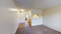 This single-story unit at 601–621 N Hayden Dr in Tucson, Arizona offers a straightforward, functional layout connecting several well-defined living spaces. The living area is finished with wall-to-wall carpet and features neutral-toned walls beneath a textured ceiling, creating a quiet, understated atmosphere. A pass-through window connects the living space directly to the kitchen, maintaining visual openness between the two areas.

The kitchen is a galley-style space laid with wood-look plank flooring and lined with warm-toned wood cabinetry above and below the counters. It includes a range with an overhead exhaust hood, a full-size refrigerator, and a dishwasher, providing essential appliances within a compact, efficient footprint. A window at the end of the kitchen allows natural light into the space.

The bedroom is generously sized with carpet flooring and features two side-by-side closets with sliding doors, offering ample built-in storage. A hallway connects the bedroom to the rest of the unit and leads to the bathroom.

The bathroom includes a vanity with an integrated sink, laminate countertop, and a large mirror above. Wood-look flooring continues here, and an adjacent utility closet provides space for a water heater and additional storage. Overall, the unit presents a clean, practical living environment suited to everyday comfort.