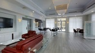 This well-appointed single-floor space offers a sophisticated and functional communal environment designed for both relaxation and social interaction. The open-plan layout is anchored by rich red leather sofas and a sleek glass coffee table, providing a comfortable lounge area that invites conversation and leisure. A set of high-top tables with modern black bar-height chairs occupies the central portion of the room, offering a versatile space for casual meetings or dining. Adjacent to the seating area, a café-style counter with a distinctive striped awning and stone accent wall adds a charming focal point, evoking a welcoming café atmosphere. Large floor-to-ceiling windows dressed in sheer white curtains flood the interior with natural light, while glass doors open directly to an outdoor terrace, seamlessly connecting the interior to the exterior. The tray ceiling, adorned with elegant pendant light fixtures and recessed lighting, adds architectural depth and a refined finishing touch. Light-toned large-format tile flooring ties the entire space together, contributing to the airy, contemporary aesthetic throughout.