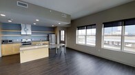 This single-floor space showcases an open-concept layout that seamlessly connects the kitchen and living area, creating a sense of spaciousness and ease of movement throughout. The kitchen is outfitted with light-toned cabinetry, a stainless steel refrigerator, and a built-in sink set into a peninsula-style counter, which also functions as a casual dining or workspace surface. An additional freestanding table adjacent to the peninsula provides extra utility within the kitchen zone. The living area is defined by rich, dark hardwood flooring that extends throughout the space, offering a warm yet modern aesthetic contrast to the neutral taupe and gray wall tones. Multiple double-hung windows fitted with black roller shades allow abundant natural light to fill the interior while offering views of the surrounding exterior. Recessed lighting in the kitchen area supplements the natural light, ensuring consistent illumination. The open floor plan, clean lines, and cohesive color palette contribute to a contemporary and functional ambiance suited for everyday living.