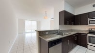 Unit 603 is a well-appointed single-floor residence characterized by a clean, contemporary design carried consistently throughout its spaces. The kitchen is a standout feature, equipped with dark espresso cabinetry, granite countertops, and a full suite of stainless steel appliances, including a refrigerator, range, and built-in microwave. A peninsula with a stainless steel sink connects the kitchen to the living and dining area, creating an open, functional flow. The living area is generously proportioned, bathed in natural light through floor-to-ceiling sliding glass doors that open to a balcony with open city views. White large-format tile flooring extends throughout the entire unit, lending a bright and cohesive aesthetic. The bedrooms are comfortably sized, each featuring windows that frame urban skyline views and allow ample natural light. Two bathrooms serve the unit; one includes a soaking tub with a tiled surround, while the other features a glass-enclosed walk-in shower. Both bathrooms are finished with granite-topped white vanities, large mirrors, and white tile flooring, maintaining the unit's consistent, modern palette throughout.