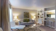 This single-floor space showcases a thoughtfully designed living area that balances comfort and contemporary style. The living room serves as the centerpiece, featuring a plush, light gray sofa accented with green and patterned throw pillows, creating a welcoming focal point. A sleek glass coffee table and modern chrome-legged accent chairs and ottoman add a sophisticated touch to the space. The room is anchored by warm, neutral-toned walls that complement the soft carpeted flooring, lending the space a cozy yet refined feel. A built-in white shelving unit lines one wall, offering both functional storage and display space for books and decorative accessories. Ambient lighting is thoughtfully layered throughout, with matching silver-based table lamps on either side of the seating area casting a warm, even glow across the room. A striking mosaic-style wall art piece serves as a tasteful focal point above the sofa, adding color and visual interest. The smooth, light-toned ceiling enhances the sense of openness, while the overall neutral palette creates a calm, versatile environment. The layout is open and efficiently arranged, making excellent use of the available space to create a functional and aesthetically pleasing living environment.