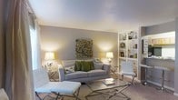 This single-floor space showcases a thoughtfully designed living area that balances comfort and contemporary style. The living room serves as the centerpiece, featuring a plush, light gray sofa accented with green and patterned throw pillows, creating a welcoming focal point. A sleek glass coffee table and modern chrome-legged accent chairs and ottoman add a sophisticated touch to the space. The room is anchored by warm, neutral-toned walls that complement the soft carpeted flooring, lending the space a cozy yet refined feel. A built-in white shelving unit lines one wall, offering both functional storage and display space for books and decorative accessories. Ambient lighting is thoughtfully layered throughout, with matching silver-based table lamps on either side of the seating area casting a warm, even glow across the room. A striking mosaic-style wall art piece serves as a tasteful focal point above the sofa, adding color and visual interest. The smooth, light-toned ceiling enhances the sense of openness, while the overall neutral palette creates a calm, versatile environment. The layout is open and efficiently arranged, making excellent use of the available space to create a functional and aesthetically pleasing living environment.