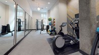 This single-level fitness facility presents a thoughtfully designed workout environment with a clean, contemporary aesthetic throughout. The main exercise area is generously proportioned, featuring light gray carpet flooring, a drop ceiling with recessed lighting, and large floor-to-ceiling mirrors framed in black that run along one wall, effectively amplifying the sense of space. Natural light filters in through windows at the far end of the room, complementing the overhead pendant lighting fixtures that add a refined touch to the space.

The workout area is equipped with a cable machine, weight bench, dumbbell rack, and an assortment of medicine balls and stability balls organized along the walls. A dedicated cardio alcove houses an elliptical and treadmill positioned against a striking herringbone wood-panel accent wall, blending light and gray-toned planks for a stylish visual contrast.

The entryway area features durable carpet tile flooring, a black-framed glass door providing sightlines into the main corridor, and a built-in stainless steel water bottle filling station with overhead shelving for towels. A collapsible hamper nearby adds practical functionality. The overall ambiance is professional, modern, and well-organized.