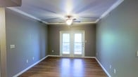 This single-story space offers a clean, well-maintained interior highlighted by warm hardwood flooring that runs throughout the main living area. The room is framed by crisp white crown molding and white baseboards, which contrast neatly against the neutral, earthy taupe-toned walls, creating a cohesive and polished aesthetic. A ceiling fan with a built-in light fixture is centrally mounted, providing both air circulation and overhead illumination. A notable feature of this space is the set of French doors centered along one wall, fitted with blinds and flanked by white trim. These doors allow a generous amount of natural light to filter into the room while offering access to what appears to be an outdoor area, such as a balcony or patio. The tray ceiling adds a subtle architectural detail that enhances the sense of height and dimension. Overall, the space presents a bright, move-in ready environment with classic finishing details that lend a timeless, functional character to the interior.
