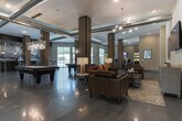 Located at 5801 Zebulon Rd in Macon, Georgia, this single-floor community clubhouse showcases a bold industrial-chic aesthetic throughout its open, well-appointed layout. Polished concrete floors reflect light across the expansive main gathering area, which is anchored by two full-sized billiard tables beneath stylish pendant and chandelier lighting. Exposed ductwork running across the high ceilings reinforces the space's contemporary industrial character, while dark-clad structural columns add architectural definition.

A standout feature is the dramatic large-scale photographic mural depicting vintage Southern Railway freight cars, which spans the length of the bar and kitchen area, serving as a striking nod to local history. The bar features granite countertops, dark cabinetry, bar-height seating, and pendant lighting, complemented by built-in cabinetry and an adjacent bookshelf with community resources.

A comfortable lounge area offers plush leather seating, an electric fireplace, a wall-mounted television, and illuminated built-in shelving — creating an inviting retreat within the larger space. Large windows along one side of the clubhouse invite natural light and provide views of the outdoor surroundings. A glass-walled fitness center is also visible from the main area, offering transparency and accessibility. Together, these zones create a versatile, amenity-rich environment designed for both relaxation and social engagement.