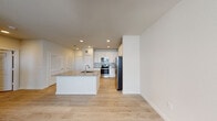 This single-floor residence showcases a clean, contemporary design throughout its open layout. The kitchen serves as the centerpiece of the living space, featuring crisp white shaker-style cabinetry extending to the ceiling, a light gray subway tile backsplash, and a full suite of stainless steel appliances including a range, built-in microwave, and French door refrigerator. A generously sized center island with a granite countertop and undermount sink provides both additional prep space and a natural gathering point. Light-toned hardwood-style flooring runs continuously throughout, enhancing the cohesive and airy feel of the space. Recessed lighting illuminates the kitchen and adjoining living area evenly. The bathroom is well-appointed with a white vanity topped with granite, a large wall mirror with overhead lighting, a wall-mounted storage cabinet, and a soaking tub with marble-style tiled surround. Additional storage is provided by a spacious walk-in closet fitted with multi-level wire shelving. The overall design palette of neutral whites and warm wood tones creates a bright, modern ambiance throughout.