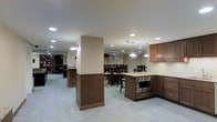 This single-floor space is designed to accommodate both food preparation and group dining in a cohesive, open-plan layout. The kitchen area is outfitted with warm medium-toned wood cabinetry, granite countertops, and stainless steel appliances, including a built-in microwave. A central structural column with matching wood paneling anchors the space, lending it a polished, intentional design aesthetic. Pendant lights hang above the kitchen counter, complementing a grid of recessed ceiling lights that illuminate the entire area evenly and brightly. The flooring throughout is large-format light grey tile, providing a clean and durable surface well-suited to high-traffic use. Adjacent to the kitchen, a spacious dining area features multiple tables and chairs arranged to seat larger groups comfortably, making the space ideal for communal meals or events. Neutral beige walls and white ceilings create a calm, neutral backdrop, while small egress windows along the far wall allow for a touch of natural light. A secondary area is visible in the background, hinting at additional functional space, possibly a lounge or storage zone. Overall, the space balances practicality with thoughtful design details, creating an environment that is both functional and inviting.