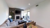 This single-floor space showcases a contemporary living area designed with clean lines and a neutral palette. The living room centers around a tiled fireplace surround set against a charcoal accent wall, above which a large wall-mounted television is positioned for an integrated entertainment focal point. Recessed lighting in the white ceiling provides consistent illumination throughout the space, complementing the natural light that filters in through roller shades on the sliding glass doors. These doors open directly onto a furnished balcony, where outdoor seating is visible, extending the living area and offering a connection to the outdoors. Light hardwood-style flooring runs throughout, contributing to the bright and airy feel of the space. Seating includes a generously sized neutral sectional sofa accented with blue throw pillows, paired with a tufted leather ottoman serving as a central coffee table. A rustic wooden coffee table and decorative ceramic vases add contrast and texture to the modern setting. A tall indoor plant near the fireplace introduces a natural element, softening the overall aesthetic and adding warmth to the contemporary design.