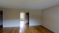 This single-floor space offers a straightforward and functional layout characterized by its warm hardwood flooring that runs throughout, adding a natural richness to the otherwise neutral interior. The main room is open and unadorned, with smooth white walls and a clean white ceiling that together create a bright, airy atmosphere. The simplicity of the space allows for versatile furniture arrangements and personalization.

An open doorway leads to an adjoining room, which benefits from a window that allows natural light to spill into the space, brightening both areas. A closet is also accessible from this secondary room, providing useful built-in storage. A baseboard heating unit is visible beneath the window, indicating a practical heating setup integrated along the perimeter of the room.

The overall ambiance is understated and move-in ready, with a neutral color palette of whites and warm wood tones forming a cohesive foundation. The hardwood floors throughout lend a sense of continuity and quality to the space, making it an adaptable environment suitable for a range of uses.