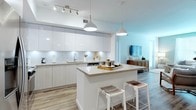 This well-designed single-floor residence blends modern aesthetics with functional living across its thoughtfully arranged spaces. The open-concept kitchen is a standout feature, showcasing high-gloss white upper cabinetry, light wood-toned lower cabinets, and a classic subway tile backsplash with under-cabinet lighting. A central island with a light quartz countertop provides additional prep space and casual seating with woven bar stools. Stainless steel appliances, including a French door refrigerator, electric range, and built-in microwave, complete the contemporary kitchen suite. Light wood-look flooring flows seamlessly throughout the unit, enhancing the sense of continuity and warmth. The living area features a soft sage green accent wall, a neutral sectional sofa, and a natural wood coffee table, creating a relaxed yet refined atmosphere. The bedroom offers a serene retreat with light gray walls, a graceful iron-frame bed, large windows dressed with white sheer curtains for natural light, and ample closet access. The bathroom is finished with a white quartz vanity countertop, a large mirror, wall sconce lighting, a deep soaking tub with subway tile surround, and a bold dark green accent wall that adds a modern, spa-like contrast. Overall, the space delivers a cohesive, contemporary living experience with clean lines and a carefully curated neutral palette.