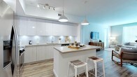 This well-designed single-floor residence blends modern aesthetics with functional living across its thoughtfully arranged spaces. The open-concept kitchen is a standout feature, showcasing high-gloss white upper cabinetry, light wood-toned lower cabinets, and a classic subway tile backsplash with under-cabinet lighting. A central island with a light quartz countertop provides additional prep space and casual seating with woven bar stools. Stainless steel appliances, including a French door refrigerator, electric range, and built-in microwave, complete the contemporary kitchen suite. Light wood-look flooring flows seamlessly throughout the unit, enhancing the sense of continuity and warmth. The living area features a soft sage green accent wall, a neutral sectional sofa, and a natural wood coffee table, creating a relaxed yet refined atmosphere. The bedroom offers a serene retreat with light gray walls, a graceful iron-frame bed, large windows dressed with white sheer curtains for natural light, and ample closet access. The bathroom is finished with a white quartz vanity countertop, a large mirror, wall sconce lighting, a deep soaking tub with subway tile surround, and a bold dark green accent wall that adds a modern, spa-like contrast. Overall, the space delivers a cohesive, contemporary living experience with clean lines and a carefully curated neutral palette.