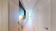 This single-floor space showcases a thoughtfully designed modern interior with a cohesive, minimalist aesthetic throughout. The kitchen is a standout feature, fitted with flat-front light wood cabinetry paired with bold teal-toned upper cabinets, a sleek grey composite countertop, an undermount stainless steel sink with a professional-style spring faucet, and under-cabinet lighting that highlights the clean lines of the workspace. Appliances are integrated seamlessly into the cabinetry for a streamlined look.

Light oak hardwood flooring runs continuously throughout the unit, lending warmth and visual continuity to every room. The living and sleeping areas are bright and airy, benefiting from floor-to-ceiling windows that frame views of the surrounding neighborhood and invite generous amounts of natural light. The crisp white walls and clean ceilings amplify the sense of openness.

The bathroom is finished with large-format neutral stone-look tiles on both the floors and walls, a frameless glass walk-in shower with a built-in bench, and warm wood-accented cabinetry. A sliding frosted glass door adds a contemporary touch to one of the rooms. Discreet panel radiators provide heating, and track lighting in the kitchen corridor ensures well-lit functionality. The overall ambiance is calm, clean, and refined.