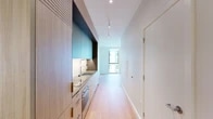 This single-floor space showcases a thoughtfully designed modern interior with a cohesive, minimalist aesthetic throughout. The kitchen is a standout feature, fitted with flat-front light wood cabinetry paired with bold teal-toned upper cabinets, a sleek grey composite countertop, an undermount stainless steel sink with a professional-style spring faucet, and under-cabinet lighting that highlights the clean lines of the workspace. Appliances are integrated seamlessly into the cabinetry for a streamlined look.

Light oak hardwood flooring runs continuously throughout the unit, lending warmth and visual continuity to every room. The living and sleeping areas are bright and airy, benefiting from floor-to-ceiling windows that frame views of the surrounding neighborhood and invite generous amounts of natural light. The crisp white walls and clean ceilings amplify the sense of openness.

The bathroom is finished with large-format neutral stone-look tiles on both the floors and walls, a frameless glass walk-in shower with a built-in bench, and warm wood-accented cabinetry. A sliding frosted glass door adds a contemporary touch to one of the rooms. Discreet panel radiators provide heating, and track lighting in the kitchen corridor ensures well-lit functionality. The overall ambiance is calm, clean, and refined.