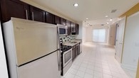 This single-floor residence offers a clean, modern aesthetic throughout its open and functional layout. The kitchen is a standout feature, showcasing rich dark espresso cabinetry that contrasts beautifully with light granite countertops and a tasteful tile backsplash in a mosaic pattern. Stainless steel appliances — including a full-size refrigerator, electric range, built-in microwave, and dishwasher — provide a sleek, cohesive look. Recessed lighting illuminates the kitchen workspace, highlighting the quality finishes. The space flows seamlessly from the kitchen into an open living area, which is bathed in natural light courtesy of large frosted glass panels or window treatments that diffuse sunlight evenly across the room. Consistent large-format white tile flooring runs throughout, tying the areas together and contributing to a bright, airy atmosphere. Track lighting in the living area adds a modern touch and allows for flexible illumination. The overall ambiance is contemporary and well-maintained, offering a comfortable and stylish environment suited for everyday living. The thoughtful design balances practicality with modern aesthetics, making the most of the available floor space.