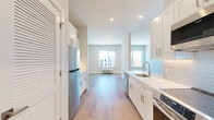 This single-floor unit showcases a contemporary design characterized by a clean, neutral color palette, light hardwood-style flooring, and recessed lighting throughout. The open-concept layout connects the kitchen and living areas seamlessly, creating a sense of spaciousness and ease of movement.

The kitchen is well-appointed with white shaker-style cabinetry, sleek quartz countertops, and a textured tile backsplash. Stainless steel appliances include a refrigerator, electric range, over-the-range microwave, and dishwasher. A louvered door provides access to additional storage, and ample cabinet space ensures strong functionality.

The living area benefits from natural light streaming through large windows that frame views of the surrounding building exterior. A wall-mounted HVAC unit provides climate control, and clean white baseboards complement the neutral walls throughout.

The bedroom is a calm, well-lit space with a large window, hardwood-style flooring, and a closet with sliding doors. The bathroom offers a tiled floor, a bathtub with shower fixtures, and a grab bar, suggesting accessibility-conscious design. An electrical panel is discreetly located in the main living area. The unit's overall aesthetic is modern, bright, and move-in ready.