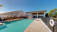 This outdoor amenity space centers around a generously sized swimming pool with crystal-clear turquoise water, accessible via built-in entry steps with stainless steel handrails. The pool is framed by a wide, paver-tiled sun deck that provides ample room for relaxation and circulation. Surrounding the pool, rows of lounge chairs and seating areas invite residents to unwind under open skies, with safety equipment such as a life ring station conveniently positioned along the perimeter. A standout feature of the space is the covered outdoor cabana structure, which boasts a warm wood-paneled ceiling, ceiling fans for year-round comfort, and a built-in bar or service counter. The cabana's open-air design, supported by clean white columns, creates a welcoming shaded retreat directly adjacent to the pool deck. Additional uncovered patio areas with umbrella-shaded tables provide further gathering spots. The entire pool area is enclosed by a secure metal fence, ensuring privacy and safety. The surrounding landscape includes mature trees and well-maintained greenery, contributing to a relaxed, resort-like ambiance. Multi-story residential buildings line the perimeter, creating an intimate courtyard feel under expansive, sun-filled skies.