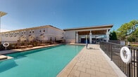 This outdoor amenity space centers around a generously sized swimming pool with crystal-clear turquoise water, accessible via built-in entry steps with stainless steel handrails. The pool is framed by a wide, paver-tiled sun deck that provides ample room for relaxation and circulation. Surrounding the pool, rows of lounge chairs and seating areas invite residents to unwind under open skies, with safety equipment such as a life ring station conveniently positioned along the perimeter. A standout feature of the space is the covered outdoor cabana structure, which boasts a warm wood-paneled ceiling, ceiling fans for year-round comfort, and a built-in bar or service counter. The cabana's open-air design, supported by clean white columns, creates a welcoming shaded retreat directly adjacent to the pool deck. Additional uncovered patio areas with umbrella-shaded tables provide further gathering spots. The entire pool area is enclosed by a secure metal fence, ensuring privacy and safety. The surrounding landscape includes mature trees and well-maintained greenery, contributing to a relaxed, resort-like ambiance. Multi-story residential buildings line the perimeter, creating an intimate courtyard feel under expansive, sun-filled skies.
