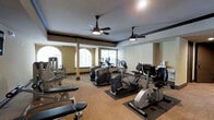 This fitness center is designed to support a full range of workout routines in a comfortable and well-appointed environment. The space features an array of cardio equipment, including elliptical trainers, stair climbers, and recumbent bikes from reputable brands such as True Fitness, all positioned on protective rubber floor mats. Strength training options are also available, with weight benches and resistance machines situated along the perimeter of the room. The layout is open and spacious, allowing for easy movement between stations. Overhead, dual ceiling fans with integrated lighting ensure consistent air circulation and a comfortable exercise environment. The walls are finished in a warm neutral tone, complemented by dark crown molding that adds a refined architectural detail. Large arched windows along one wall admit generous natural light, while blinds offer adjustable privacy and glare control. Structural columns with dark bases punctuate the space, contributing to the room's overall design character. The flooring is a durable, textured carpet suitable for a high-traffic fitness setting. Overall, the fitness center offers a practical and well-maintained space ideal for daily exercise.