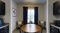 Located at 111 Springview Ln in Summerville, South Carolina, this single-floor space offers a clean, well-appointed interior designed with both comfort and style in mind. The featured seating area is a standout element of the space, centered around a warm-toned round wooden table surrounded by upholstered accent chairs in a sophisticated charcoal gray, each adorned with gold decorative pillows that add a refined touch. Floor-to-ceiling navy blue curtains frame a large window fitted with horizontal blinds, allowing for adjustable natural light while contributing to the room's polished aesthetic. A geometric gold centerpiece sits atop the table, complementing the gold-accented wall art that adds a contemporary artistic flair to the neutral-toned walls. The flooring is covered in plush carpeting, enhancing the room's warm and inviting ambiance. The overall design palette — combining navy, gold, and gray — creates a cohesive and tasteful atmosphere that feels both functional and elegantly curated. Natural light filters through the window, offering a connection to the outdoors while maintaining a comfortable, enclosed feel throughout the space.