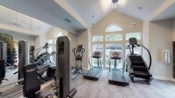 The Grand on Memorial fitness center is a thoughtfully designed, single-floor facility built to support a comprehensive workout experience. The space is anchored by soaring vaulted ceilings and floor-to-ceiling windows that flood the cardio area with natural light and offer views of the surrounding greenery, creating an open and energizing atmosphere. The cardio section houses multiple treadmills, elliptical trainers, a stair climber, and a recumbent bike, all arranged to maximize space and usability. Full-length mirrors line the walls, visually expanding the room and allowing users to monitor their form during exercise. The strength training area is equally well-appointed, featuring a cable machine, a dumbbell rack with a full range of weights, and a medicine ball tower. A striking nature-themed mural covers one wall, adding a vibrant, outdoor-inspired aesthetic to the interior. Motivational quotes are displayed throughout, complementing the branded "The Grand on Memorial" signage and teal accent wall graphics. A water station is conveniently located near the entrance. Durable, easy-to-maintain flooring ties the space together, while recessed lighting and ceiling fans ensure comfort during any workout. A glass-panel door provides direct access to the outdoor area.