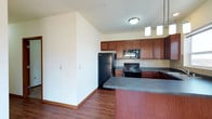 This single-floor space showcases a well-appointed kitchen as its centerpiece, designed for both functionality and style. The kitchen features rich, medium-toned wood cabinetry that extends to the upper walls, providing generous storage capacity. Dark laminate countertops run throughout, offering ample workspace, and complement the suite of black appliances, which includes a French door refrigerator, a gas range, and an over-the-range microwave. A peninsula with a matching dark countertop extends outward, creating a convenient breakfast bar or additional prep area, and is illuminated by two pendant lights with clean, cylindrical shades suspended from above. A tile backsplash accents the cooking area, adding visual texture behind the range. The flooring throughout is a warm hardwood-style surface that anchors the space and enhances its welcoming ambiance. Flush-mount ceiling lighting provides consistent illumination across the open layout. The open-concept design connects the kitchen to the adjoining living area, separated by a low partial wall, allowing the space to feel expansive and cohesive throughout.