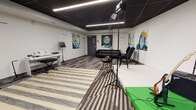 This single-floor creative studio is thoughtfully designed to support a wide range of music, recording, and multimedia production activities. The space features a striking black ceiling with sleek linear LED lighting strips that cast clean, even illumination across the room. A bold striped area rug anchors the main open floor, which flows seamlessly between distinct functional zones.

One section serves as a performance and recording area, equipped with a raised green screen platform, professional lighting stands, microphone stands, and an electric guitar with amplifier. Adjacent to this is a dedicated acoustic wall treated with sculptural three-dimensional black panels, providing sound dampening ideal for recording and rehearsal. A keyboard workstation with music stand and a small amplifier occupy another corner, offering a self-contained music practice setup.

The lounge area features a deep brown channeled leather sofa and an accent chair, creating a comfortable space for artists and collaborators to relax between sessions. Throughout the studio, large-scale colorful portrait murals of iconic music figures adorn the white walls, adding artistic energy and cultural depth to the environment. The dark ceiling treatment, acoustic paneling, professional-grade equipment, and curated artwork combine to create an inspiring, functional space purpose-built for creative production.