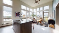 This single-floor residence is defined by its striking architectural design, most notably the soaring double-height windows that wrap around the open-concept living and kitchen area, flooding the space with abundant natural light and framing expansive panoramic city and skyline views. A ceiling fan anchors the lofty ceiling above the living room, which features a bold teal accent wall, light hardwood-style flooring, and generous proportions ideal for comfortable living and entertaining.

The kitchen is thoughtfully designed with rich dark walnut cabinetry, a herringbone tile backsplash, stainless steel appliances, and a large central island finished with a grey stone countertop and bar seating — creating a seamless connection to the living area. A dedicated hallway leads to the bedroom, which enjoys its own large windows with sweeping city views, a walk-in closet, and a clean, serene ambiance. The bathroom features white subway tile surrounding a soaking tub, maintaining the space's understated modern aesthetic. A dedicated in-unit laundry closet with a stacked washer and dryer adds practical convenience throughout.